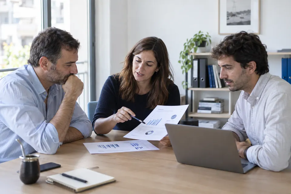 Tres personas revisando gráficos y una computadora en una oficina luminosa